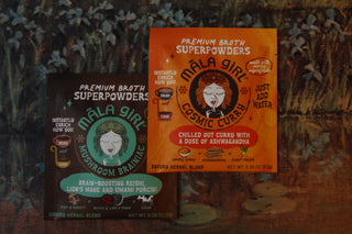 Individual broth packets for Mala Girl's Cosmic Curry and Mushroom Brainiac shown against a painting.