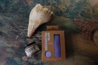 Lavender satin pillowcase shown with magnesium body butter and a seashell on top of a painting.